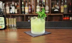 Ein grüner Tequila-Cocktail mit Minze und Eis auf einem Holztresen, im Hintergrund Regale mit Flaschen.