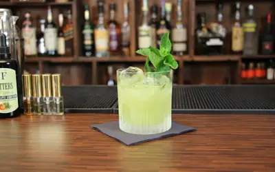 Ein grüner Tequila-Cocktail mit Minze und Eis auf einem Holztresen, im Hintergrund Regale mit Flaschen.