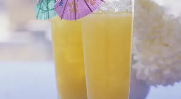 Zwei Gläser mit gelbem Saft und Eis, dekoriert mit kleinen bunten Schirmchen.