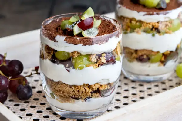 Dessert im Glas mit Schichten aus Creme, Keksbröseln, grünen und roten Trauben, garniert mit Kakaopulver.