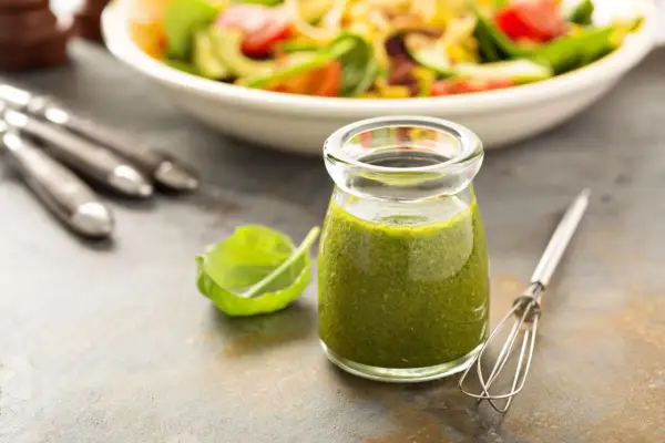 Grünes Dressing in einem Glas mit Salat und Kräutern im Hintergrund.