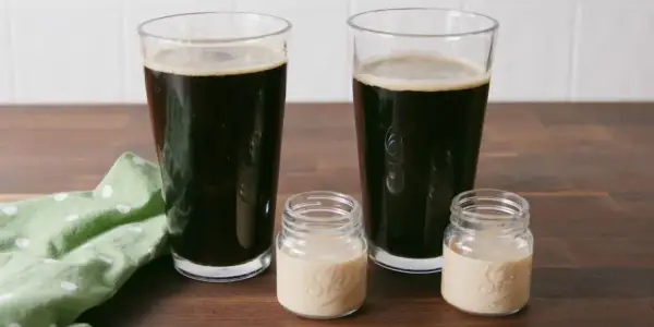 Zwei Gläser dunkles Bier und zwei kleine Gläser mit cremiger Flüssigkeit auf einem Holztisch.