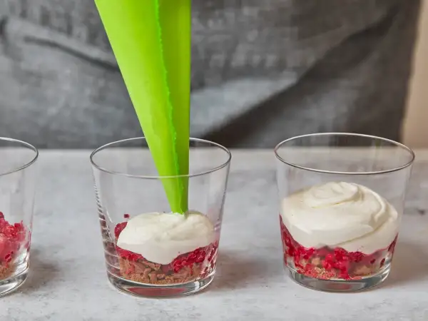 Cremige Masse wird mit Spritzbeutel in Gläser mit Himbeeren und Spekulatius-Bröseln gefüllt.
