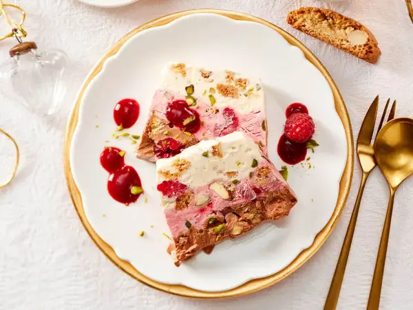 Gefrorener Kuchen mit Beeren, Nüssen und Schokolade auf einem Teller mit Sauce und Löffel.